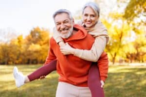 Happy mature couple enjoying piggyback ride in park smiling brightly at Dental Services at Westlake Village Dental