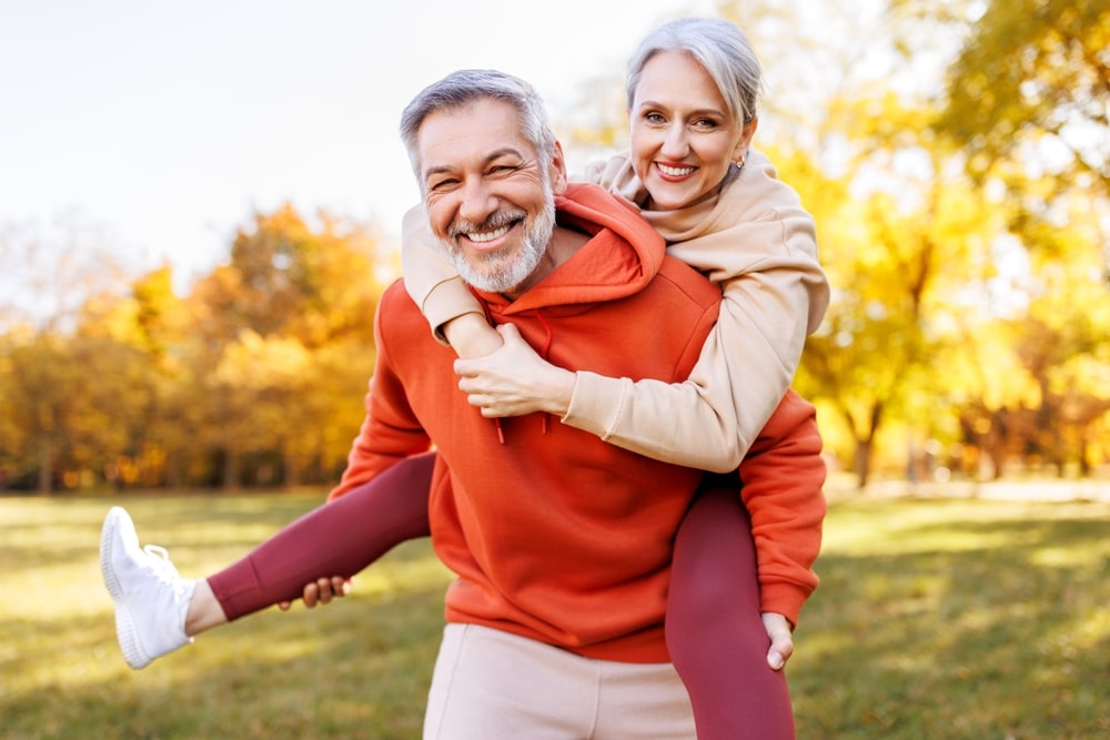 Happy mature couple enjoying piggyback ride in park smiling brightly at Dental Services at Westlake Village Dental
